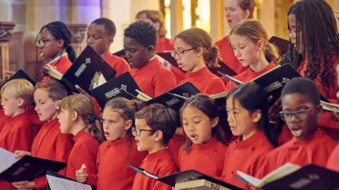 A group of children in red and white chorister frocks, singing from black books 