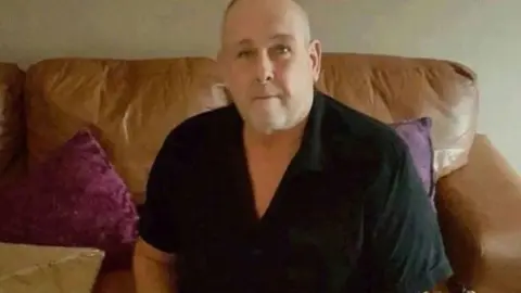 Steve Dymond/Facebook Steve Dymond looks directly at the camera, he is a sat on a brown leather sofa which has deep purple cushions on it. He wears a short sleeved black polo top