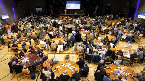 A bird's eye view of Mote Hall inside Maidstone Leisure Centre. A large number of people are sitting at circular tables.