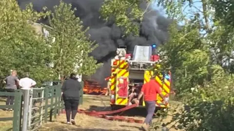 A fire engine with a long red hose coming out of the back is parked in front of large dark grey plumes of smoke, with orange flames visible. The setting is rural, with several trees nearby and a large building just visible behind trees near the fire.