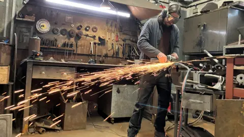 Olly Osborne, wearing goggles, is operating a metal grinder. Sparks fly across the room spectacularly. Behind him is a rack of metal-working tools and other equipment used to restore ovens.