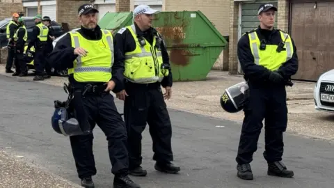 Three police officers are pictured standing in a road. They are wearing yellow vests and all have caps on