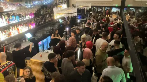 Alex Osborne/BBC Picture of a packed out bar, with rows of partygoers, some wearing colourful wigs and Santa hats. The bar is decorated with chandelier-type lighting, fairy lights and brightly coloured alcoholic bottles behind the bar. 