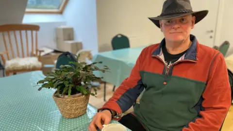 A man with a hat in a green and orange top sits with an almost empty cup of coffee in front of him. There is a plant beside him and a window behind.