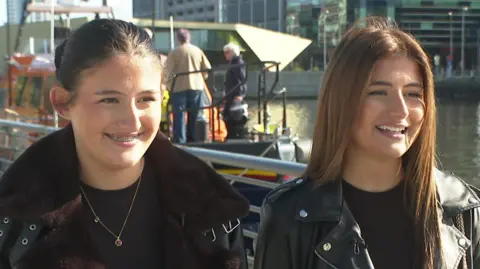 Twins Lola and Evie, both wearing dark jackets over dark tops, seen at Salford Quays with an orange RNLI lifeboat in the background