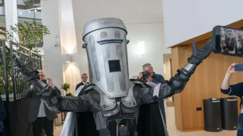 PA Media Count Binface - a man dressed in a grey space costume with a cape on the back and a dustbin for the head. He has his arms outstretched in the air while inside a venue, with a number of photographers taking pictures of him.