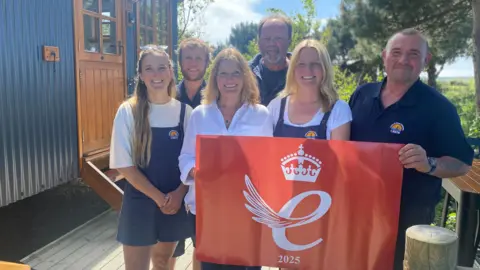 A wide shot of the Barnes family, and campsite staff. There are six of them, smiling and holding a banner with the logo for the King's Award on.