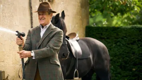 Man in tweed jacket holds a hose and is front of a horse