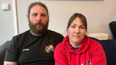 BBC Layton Jones (left) with partner Gemma Baker (right), they both sit on a sofa. Gemma wears a red pull over hoodie and looks at the camera. She has brown hair with sweeping bangs and the rest pulled back. Mr Jones has a long light brown beard and long hair parted in the middle tied back.