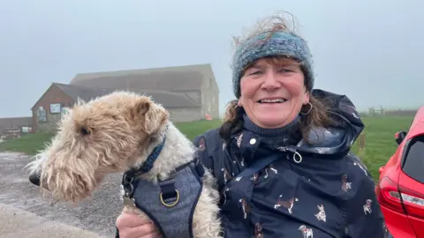 George Carden/BBC Jenny Hazelby standing in the car park holding her dog and smiling at the camera wearing a navy blue raincoat with cartoon dogs on it