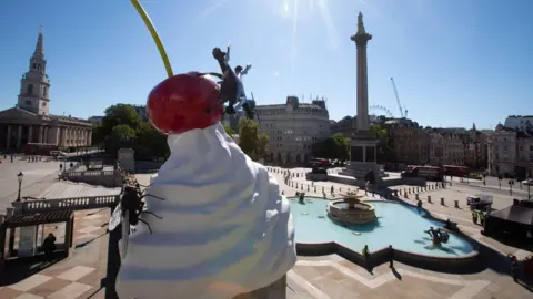 PA Media An artwork entitled THE END by Heather Phillipson, as it is unveiled on Trafalgar Square's Fourth Plinth, in London. The sculpture depicts a giant ice cream in a cone with a cherry on top and a fly on the left. Trafalgar Square is in the background.