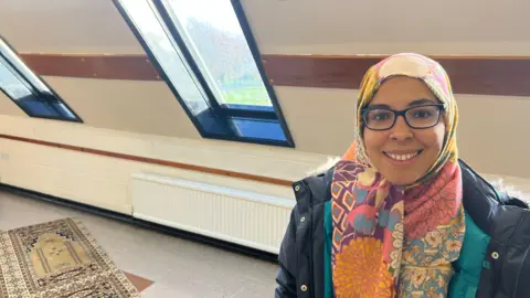 BBC/RICHARD EDWARDS Somaya Abdel-Aal - at Selby Friday prayers - is smiling and looking straight into the camera. She has a patterned headscarf, spectacles and is wearing a blue parka jacket