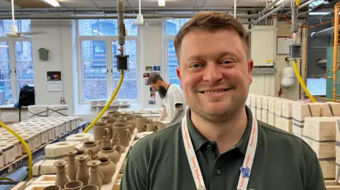 A man smiling looking directly at the camera. He has ashy brown hair and is wearing a green polo shirt. He is stood in a factory with various bits of earthenware yet to be fired.