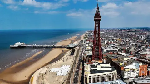 A general aerial view of Blackpool showing the tower and the town to the right, and the beach to the left with central pier. The sky is blue with a few white clouds.