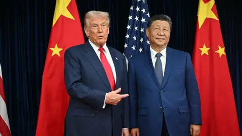 Donald Trump (L) and Xi Jinping (R), both wearing blue suits, stand between two Chinese flags, with a US flag between them. Trump is pointing towards Xi with his right hand.
