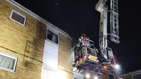 HIWFRS Firefighter standing on a ladder platform positioned at the window of a flat - it is a cream brick building with white windows.