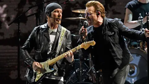 Getty Images The Edge is onstage with Bono. He's wearing a black leather jacket and playing a guitar. Bono is singing into a mic.