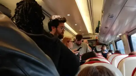 The walkway of a train carriage is filled with people standing. The tops of people's heads are visible on every seat. 
