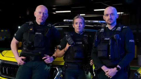 BBC/ Third Street Studios Three police officers looking at the camera. All are wearing police uniform that is black and dark green. The one on the left is a bald man, in the middle is a blonde woman and on the right is a bald man with dark beard.