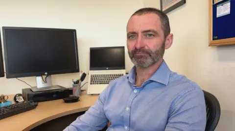 Tobin Cook sat at his desk, there is a monitor and a laptop behind him. He has short dark hair and a beard, and is wearing a blue button-up shirt.