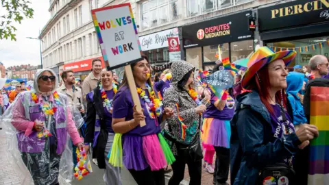 A parade of people dressed in bright, rainbow colours walk down a high street. One woman is holding a sign that says "Wolves at Work".