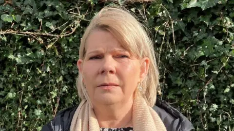 Ms Boyle has shoulder length blonde hair and is wearing a cream scarf, black patterned top and black coat as she stares at the camera. 