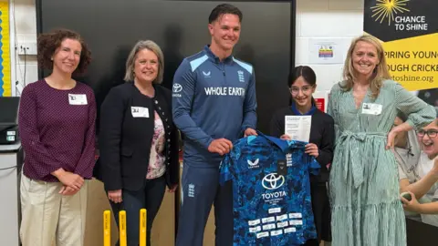 Sonny Baker, wearing England kit, presents schoolgirl Ava with a blue England shirt signed by the team. Stood next to them are three female dignitaries, including Oxford East MP Anneliese Dodds.