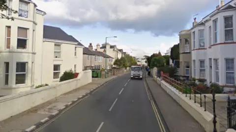 A straight road with pavements either side and a truck driving towards the camera on the right. Terraced houses line both sides of the road.