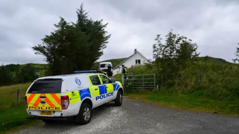 PA Media A Police car outside a house on Skye. The police car is long and white with orange and yellow striped on the back and the word "police" in large orange letters above the number plate. On the side is the blue Police Scotland logo above yellow and blue squares which cover the lower part of the car. The word "police" is on the side of the doors in blue. The house in the background is surrounded by greenery and is white with a dark roof. At least three police officers can be seen behind the vehicles and a white van is also parked outside the house.