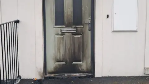 A green door with black fire marks over most of it. There is a large number in the middle of the door. The walls of the house are cream.