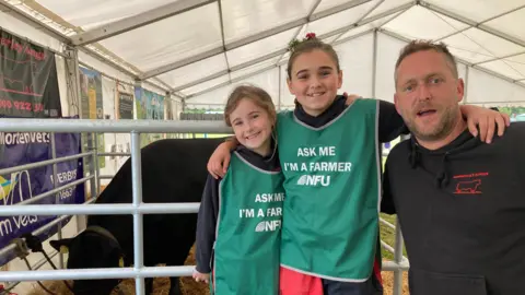 Left to right: Lily, seven, Evie, 10, who are wearing green bibs which have the words 'Ask Me I'm a Farmer NFU' (National Farmers Union)' and their father Ian at the Royal Cheshire Show. Their bull, Evolution, is in the background eating feed in a marquee at the 2026 Royal Cheshire Show. The
