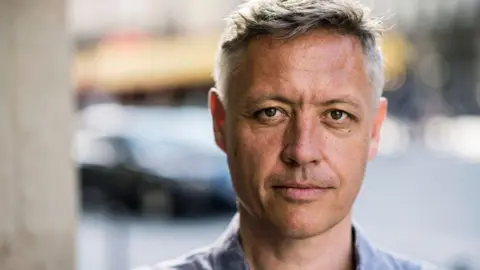 Alexandre Isard Headshot of Robert Thorogood. He is standing outside and has dark hair. He is wearing a blue shirt.