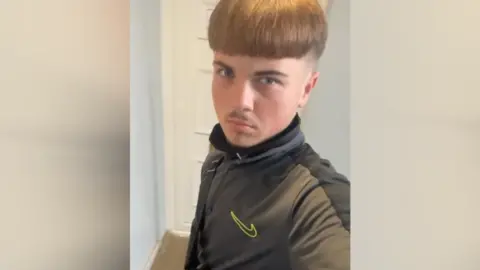 Aidan Tottman is taking a selfie with brown hair brushed to the front of his face and shaved around the side of his head. He is wearing a Nike jumper and is stood in front of a white door.