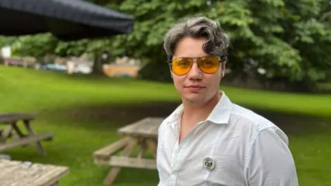 A man in a white, collared shirt is standing in a pub garden with trees and picnic benches behind him. He's wearing light yellow sunglasses and has short, light brown hair.