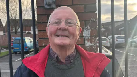 A man wearing glasses and a navy jacket with a red lining stands smiling in front of a metal fence and brick pillar, with parked cars and houses visible in the background on a bright day.