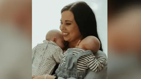 The photo shows a woman with brown hair holding two newborn babies. She is smiling and is looking to the left of the photo. One baby is wearing a flowery baby grow, and the other is wearing dungarees and a striped top. They both have their heads on the woman's shoulders. 