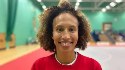 BBC Serena Kersten pictured at an indoor netball court wearing a red t-shirt. She has brown eyes and brown curly hair. 
