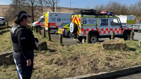 A police officer stands by some police tape and overlooks a car park filled with  mountain rescue vehicles which is acting as a base for the search.