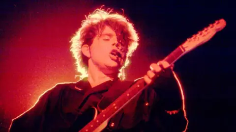 Tom Bailey plays a black electric guitar on a dark concert stage under intense red and orange lighting. Backlighting creates a bright halo around his hair during a live performance from the band's 1980s era at The Aragon Ballroom in Chicago, Illinois, on 27 April 1984.