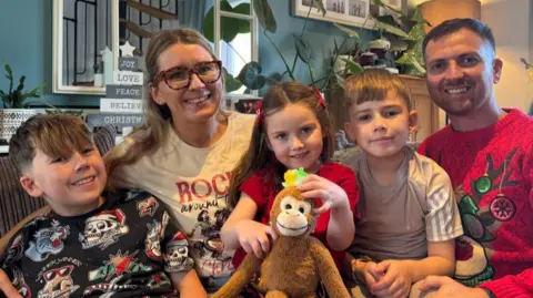 Chris McHugh/BBC A young family of five poses for a photo cuddled up on a sofa together. The two parents are both wearing Christmas jumpers and their children, two young boys and a girl, are all sat on their parents laps smiling for the camera. The young girl is holding a cuddy toy monkey.