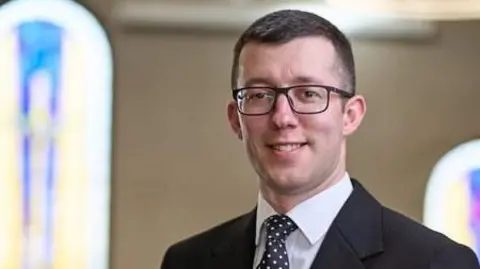 Ronnie Thompson Funeral Directors David Jess has short dark hair with black rectangular glasses, a black blazer, white shirt and black spotted tie. 