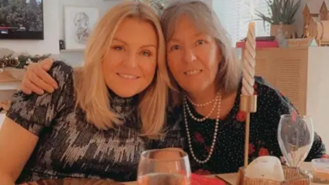 Two women sit at a table with their arms around each other. Both are smiling and looking into the camera. Empty glasses and candles can be seen in front of them.