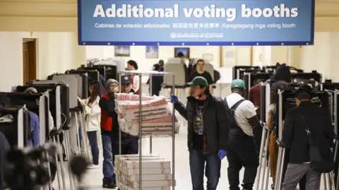 AFP via Getty Images People vote at rows of electronic voting booths inside a busy polling location. In the centre, election workers push a cart loaded with trays of ballot papers and boxes beneath a large blue sign reading “Additional voting booths” in multiple languages. The hall is lined with voting stations on both sides, with voters and staff moving through the room.