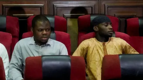 Close up of two of the men sentenced to death sitting on red and black chairs in court.