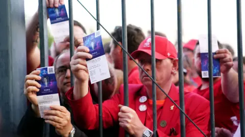 PA Media Liverpool fans clad in red shirts hold up their tickets to the Champions League 2022 final match in Paris from behind an iron gate