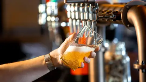 A close-up of an unrecognisable person's hand holding a pint glass at an angle being filled with beer. The beer is golden in colour, creating foam and bubbles.