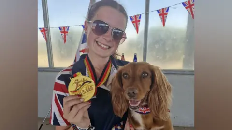 Gavin Taylor, wearing sunglasses, with her hair tied back, holding a dog, and a gold medal. Flags are behind her. She is wearing a Team GB top. 