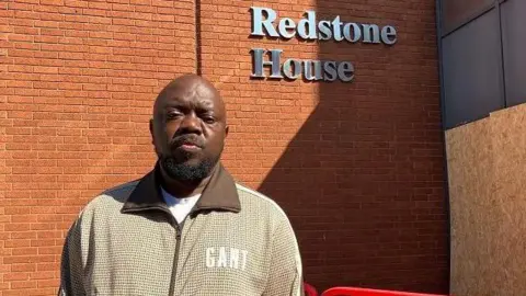 Henry Godfrey-Evans A man with short dark hair and a beard, wearing a beige zip-up jacket. He is standing in front of a brick wall with "Redstone House" in silver lettering on it. There is wooden boarding around the entrance porch, and a low red temporary fence in front of the entrance.