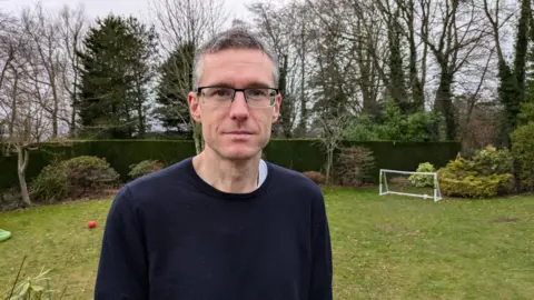 Mark Church Mark Church is standing in a garden with a hedge behind him. He is a man in his 40s with glasses and wearing a blue jumper.