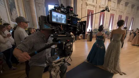 Courtesy of Netflix Film crew member operating a large camera rig inside an opulent ballroom with tall columns, draped curtains, and chandeliers.